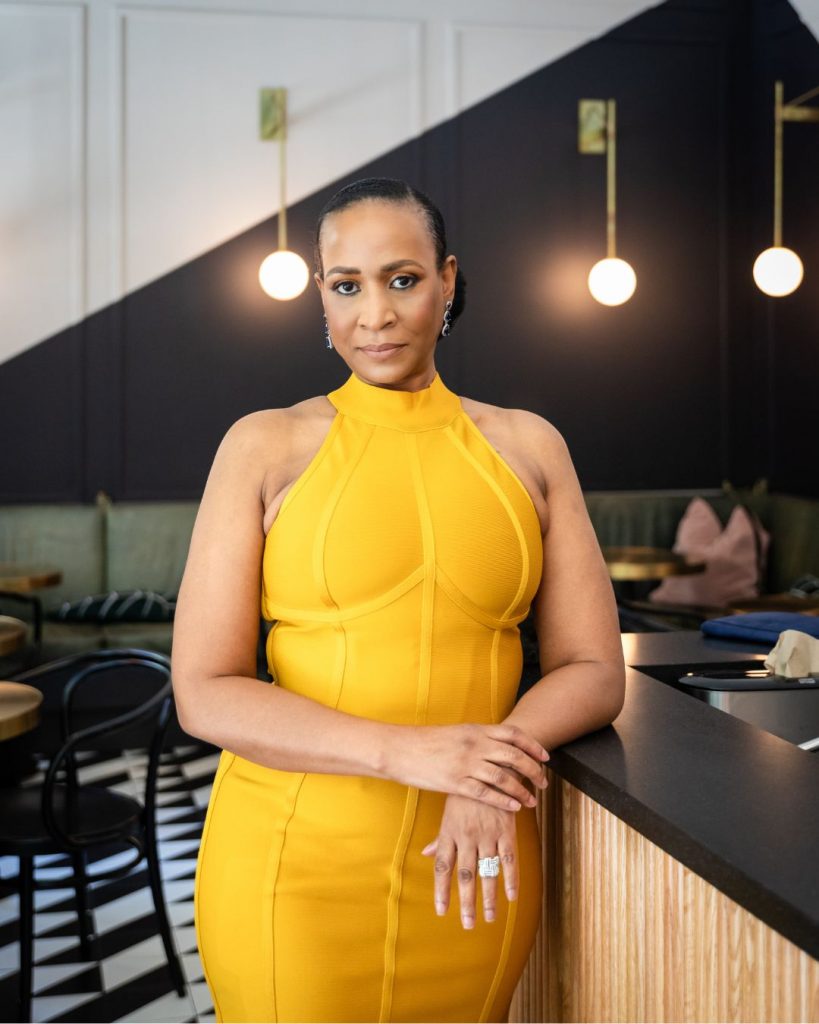 Close-up portrait of Lana Reid in a yellow dress, leaning against a counter beneath warm modern lighting.