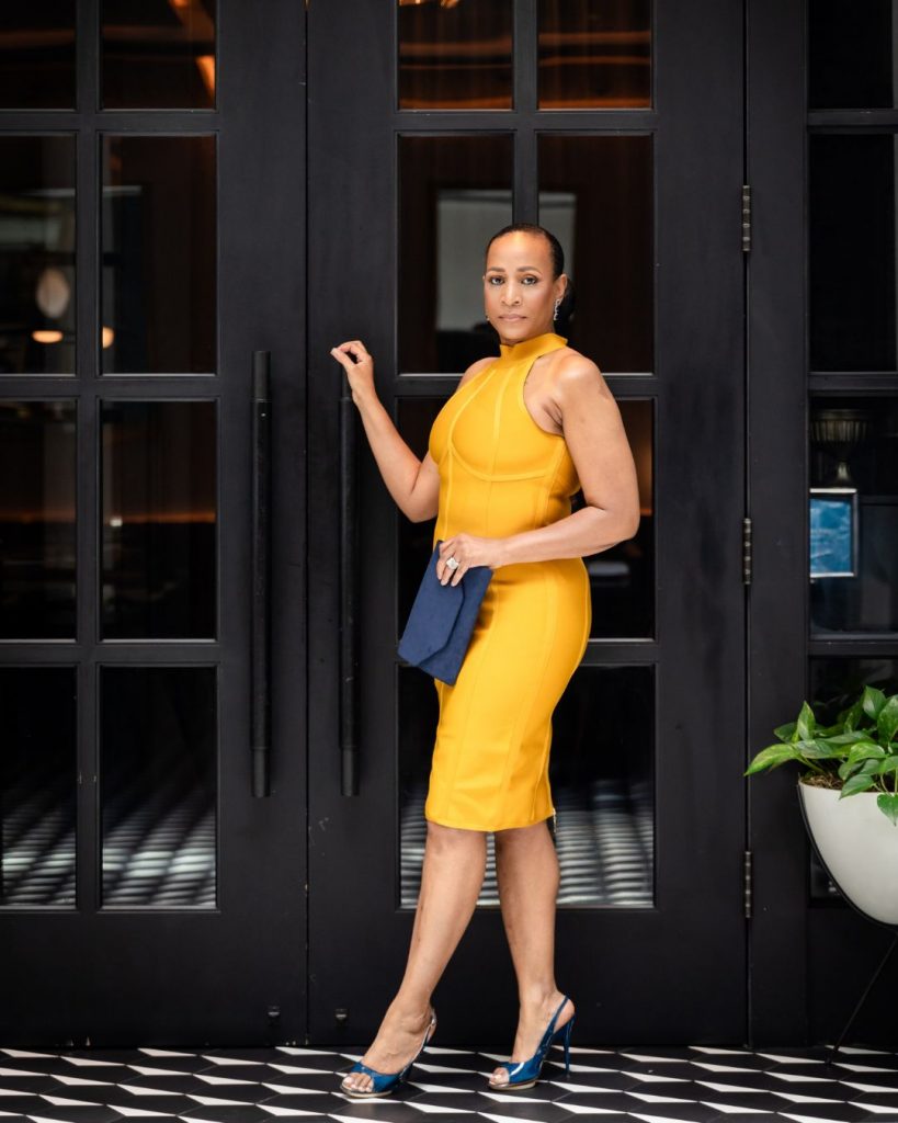 Lana Reid standing confidently in a yellow dress in front of elegant black double doors during her 55th birthday photoshoot.