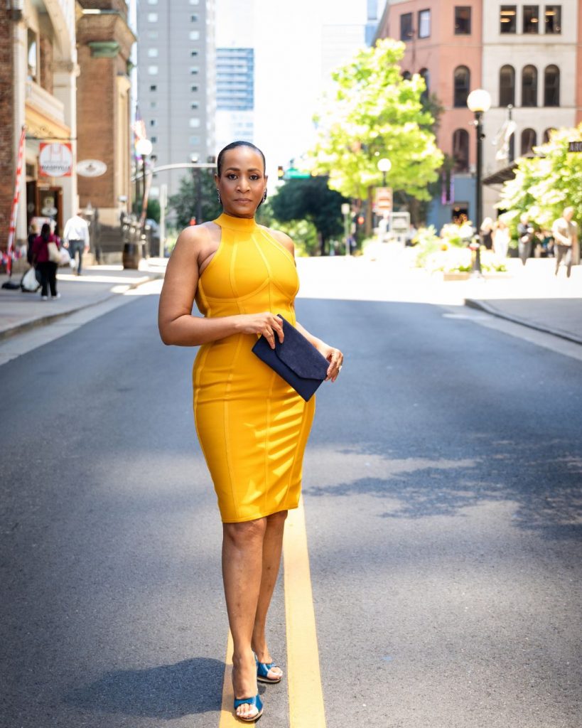Lana Reid standing in the middle of a downtown street in a yellow dress, holding a navy clutch during her 55th birthday photoshoot.