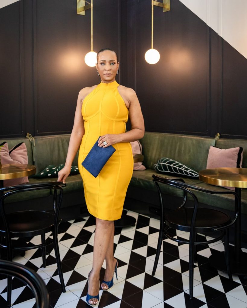 Lana Reid standing in a modern lounge space wearing a yellow dress, holding a navy clutch during her 55th birthday photoshoot.
