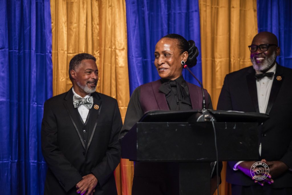 Lana Reid speaking at the podium during the 2025 Humanitarian Award ceremony with members of the Eta Nu Nu Chapter of Omega Psi Phi Fraternity standing behind her.