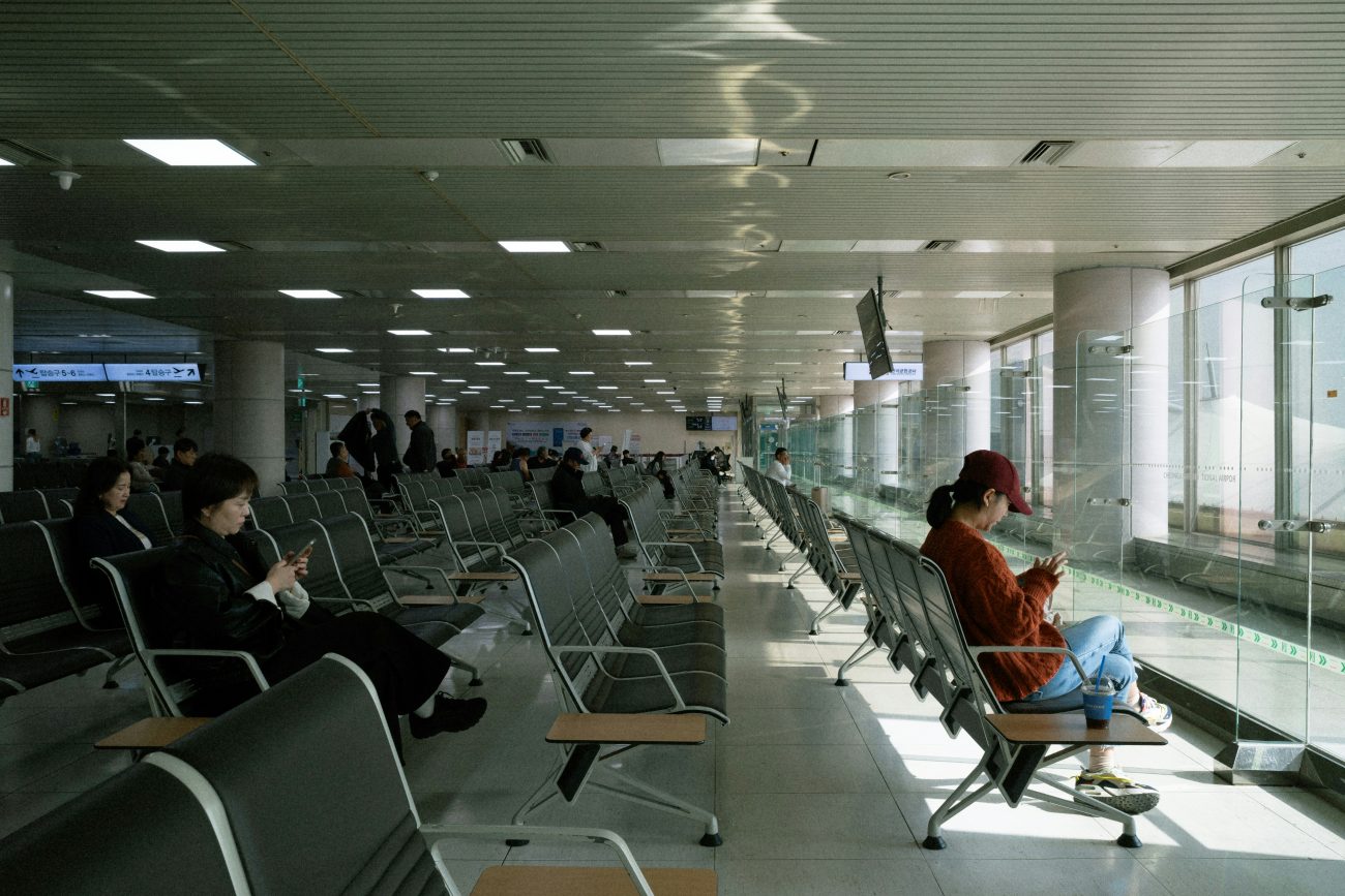 Travelers seated in an airport boarding area, most looking down at their phones while waiting for their flights.
